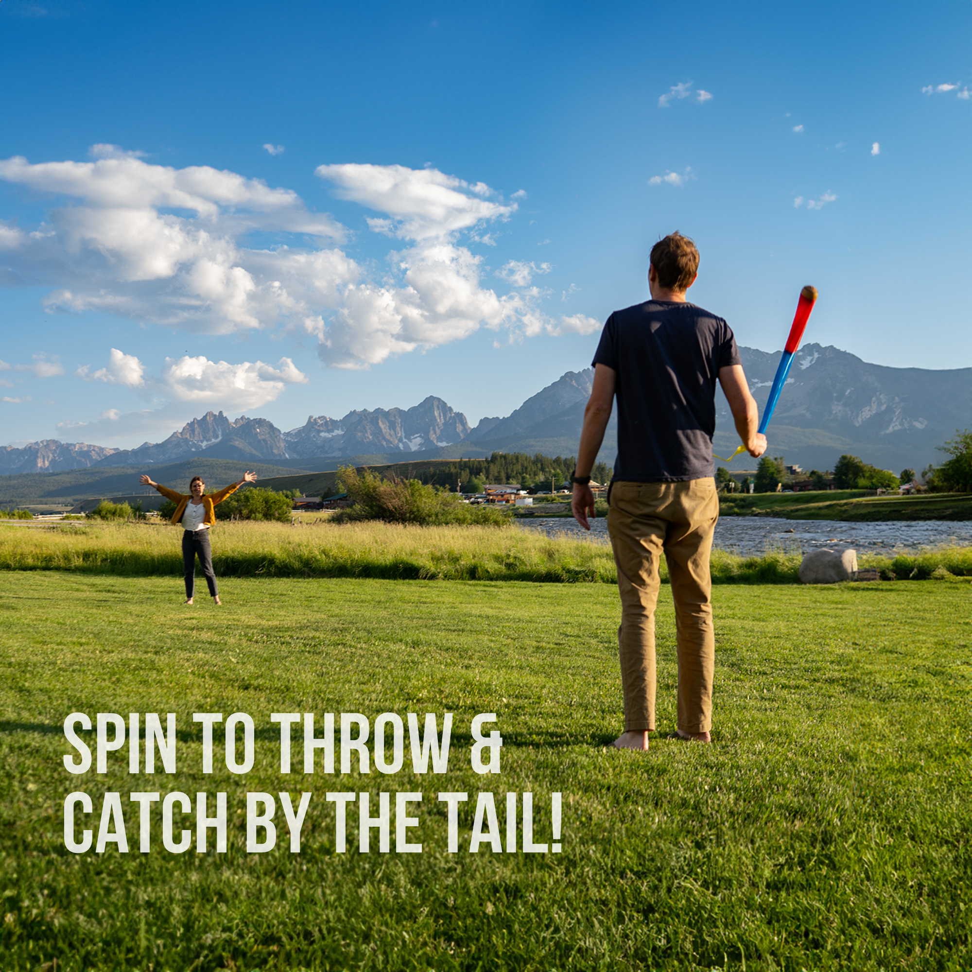A man stands on grass holding The Original Foxtail Sport, ready for outdoor play, while another person waits with arms wide. Mountains, trees, and a lake are in the background. Text reads: Spin to throw & catch by the tail!.